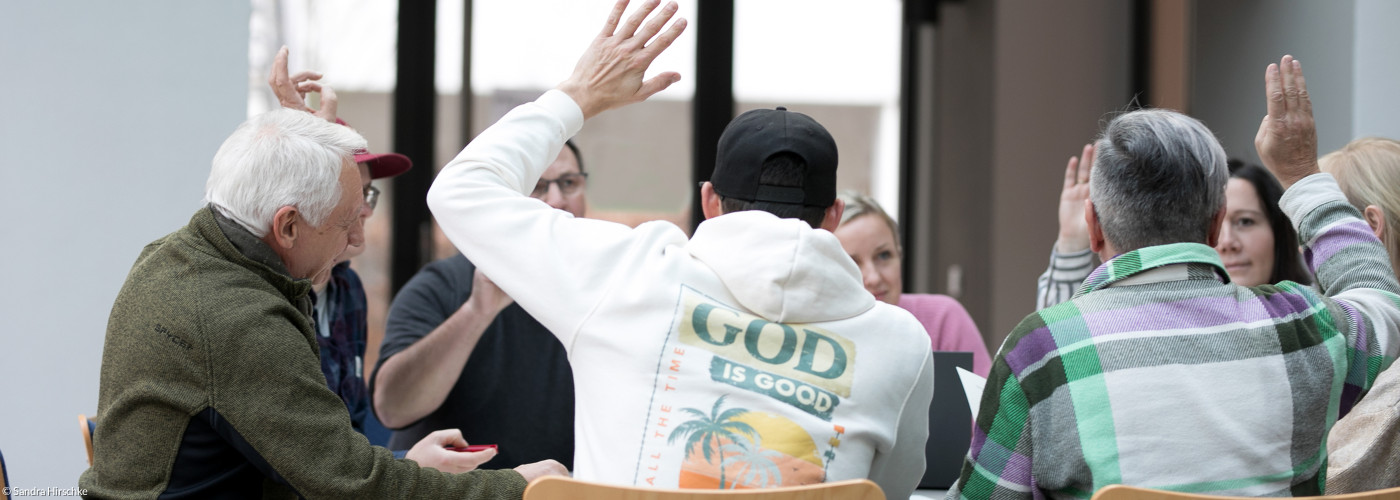 Einige Menschen sitzen am Tisch und heben die Hand bei einer Abstimmung